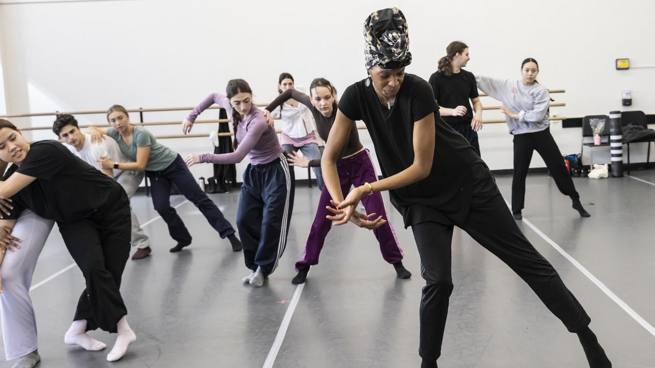 As part of a dance class, students practicing movements together. The central figure, a teacher, leads with dynamic energy, her body leaning forward in a guiding pose. She seems to be demonstrating a move, her hands outstretched as if to weave through the air, while her students, wearing various casual dance attire, mimic her stance with varying degrees of focus and precision. The mirrored wall reflects the concentrated ambiance of the room, creating an immersive environment of learning and movement.